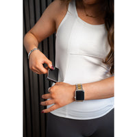 Woman wearing a white Mude Tank top and black pants, holding an insulin pump with a blurred background.