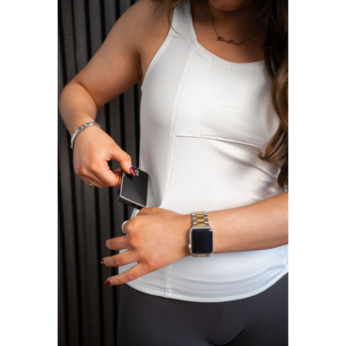 Woman wearing a white Mude Tank top and black pants, holding an insulin pump with a blurred background.