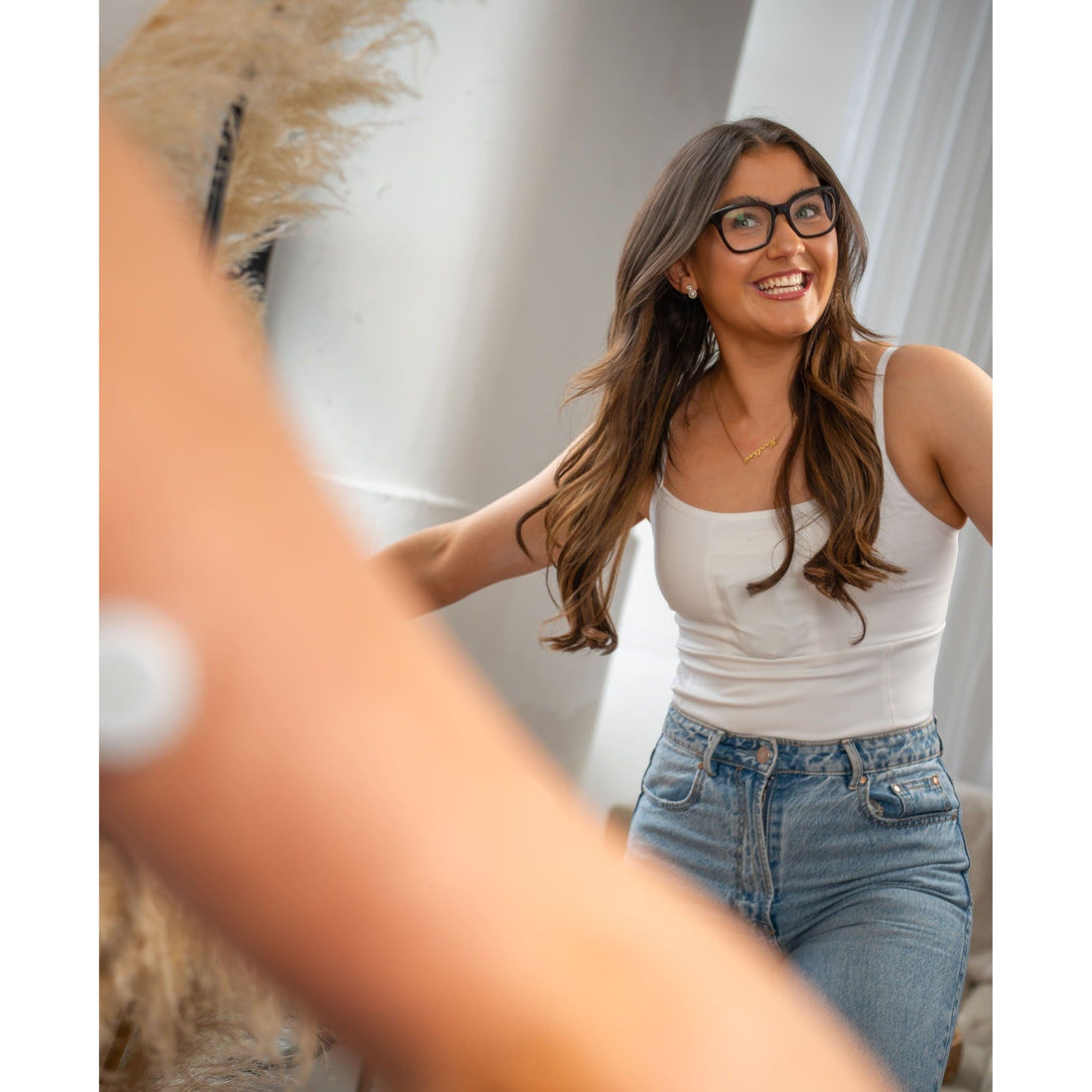 Woman wearing a white Mude Camisole top and blue jeans, smiling in a room with a couch and decorative items.