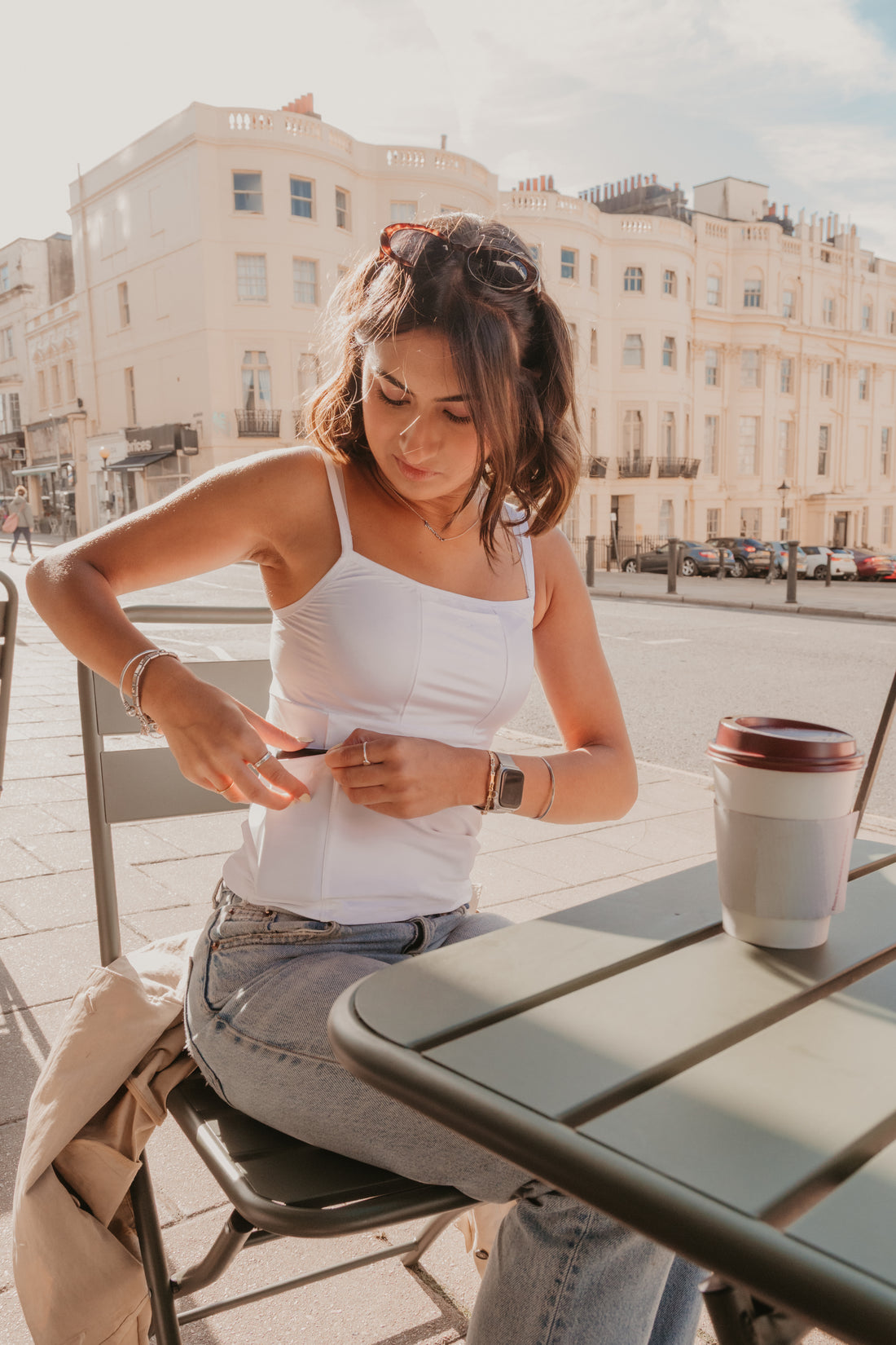 Woman sitting at an outdoor table with a coffee cup, surrounded by buildings and street wearing a white Mude Camisole.