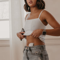 Woman wearing a white Mude Camisole top and grey jeans indoors with an Omnipod on show on her stomach.