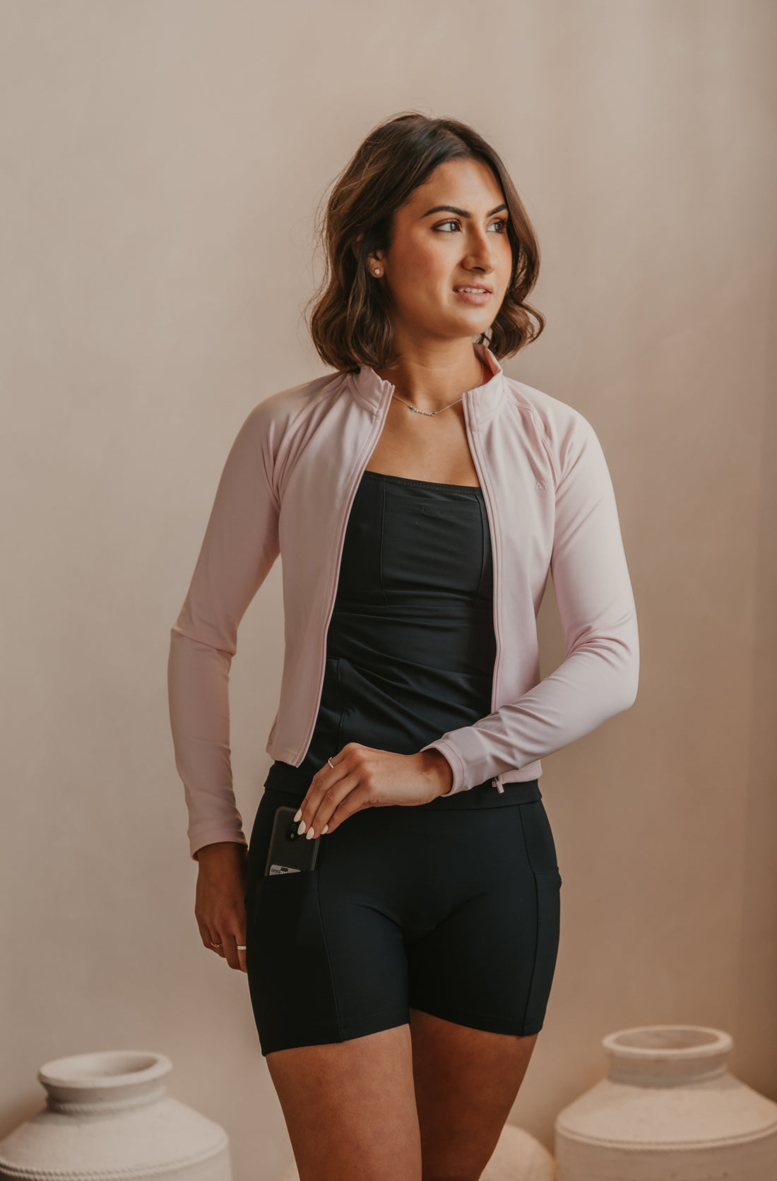 Woman wearing a light pink cardigan over a black Mude Tank and black Mude Shorts, standing against a beige wall with white vases.